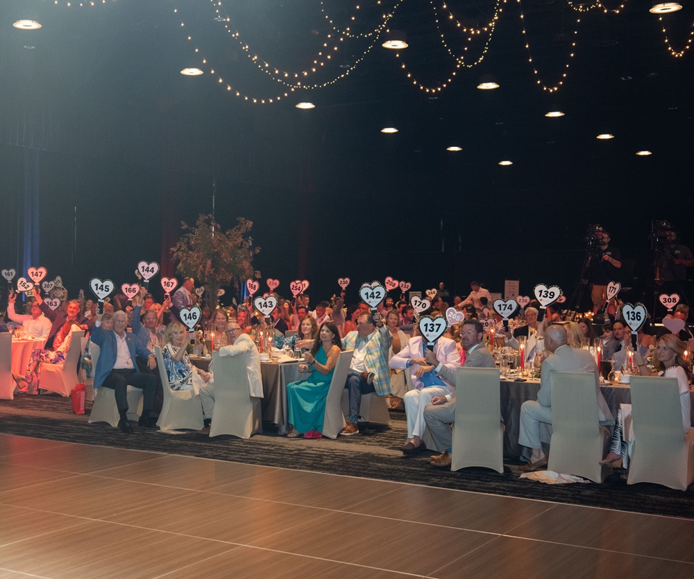 Crowded ballroom with many people holding up bidder paddles to make Open Your Heart donations.